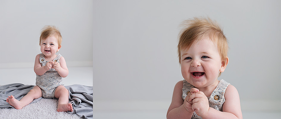 7 month old baby boy sitting on a grey rug clasping his hands and smiling