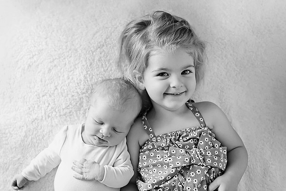 Black and white photo of a newborn baby sleeping next to a toddler smiling